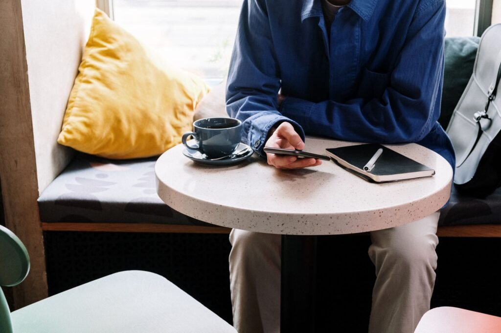 Person in blue shirt using smartphone at cafe with coffee and notepad.