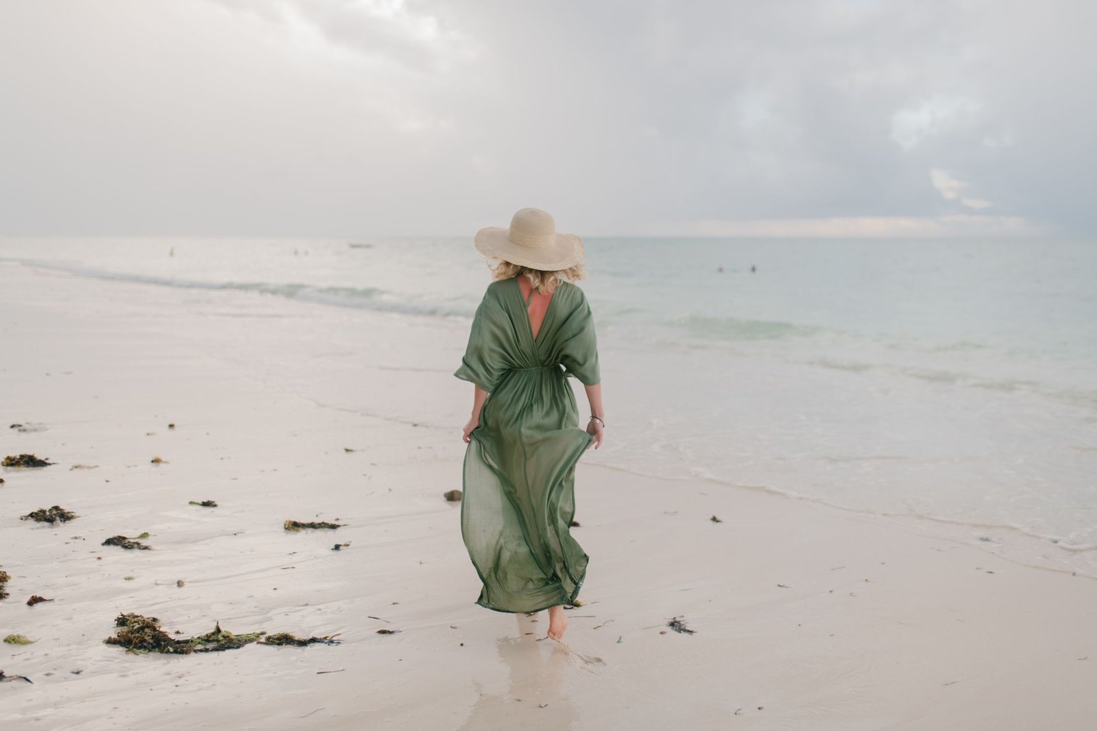 Full body back view of unrecognizable barefooted female tourist in thin dress and straw hat strolling on sandy beach towards