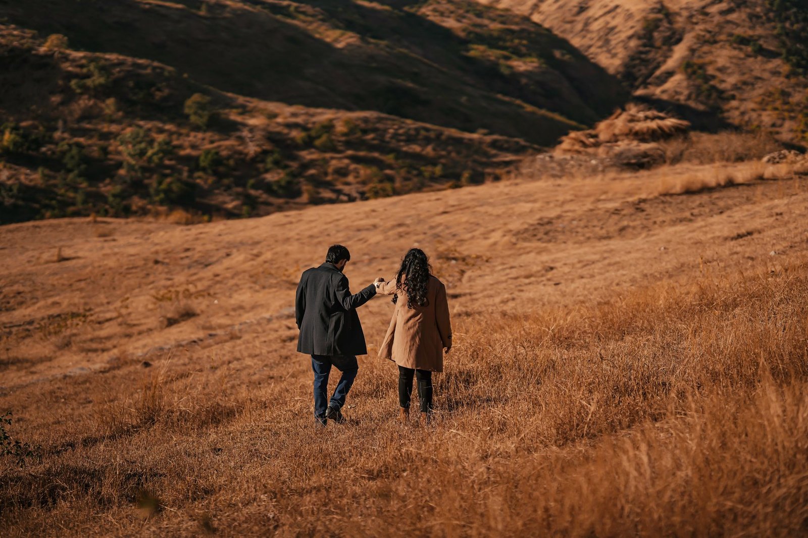 A couple walking hand in hand through a dry, hilly landscape during autumn, symbolizing romance and adventure.