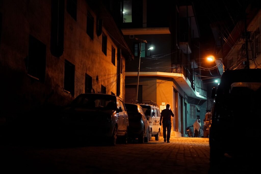 A dimly lit urban alley at night with silhouetted people walking amidst parked cars under streetlights.