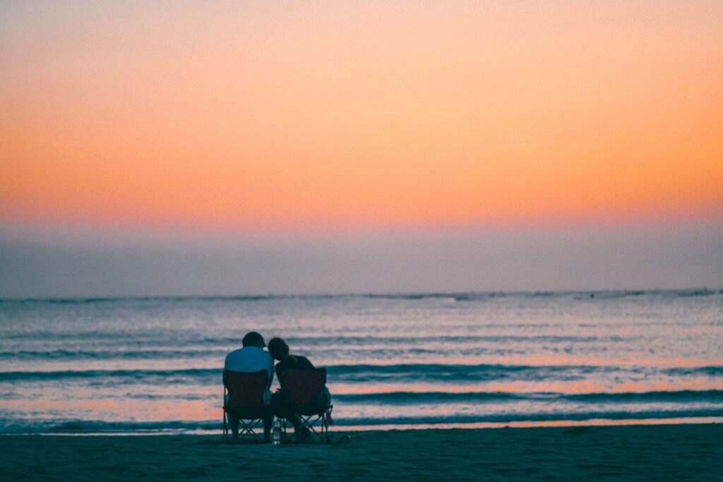 A couple sits in chairs on the beach, watching a beautiful sunset over the ocean.