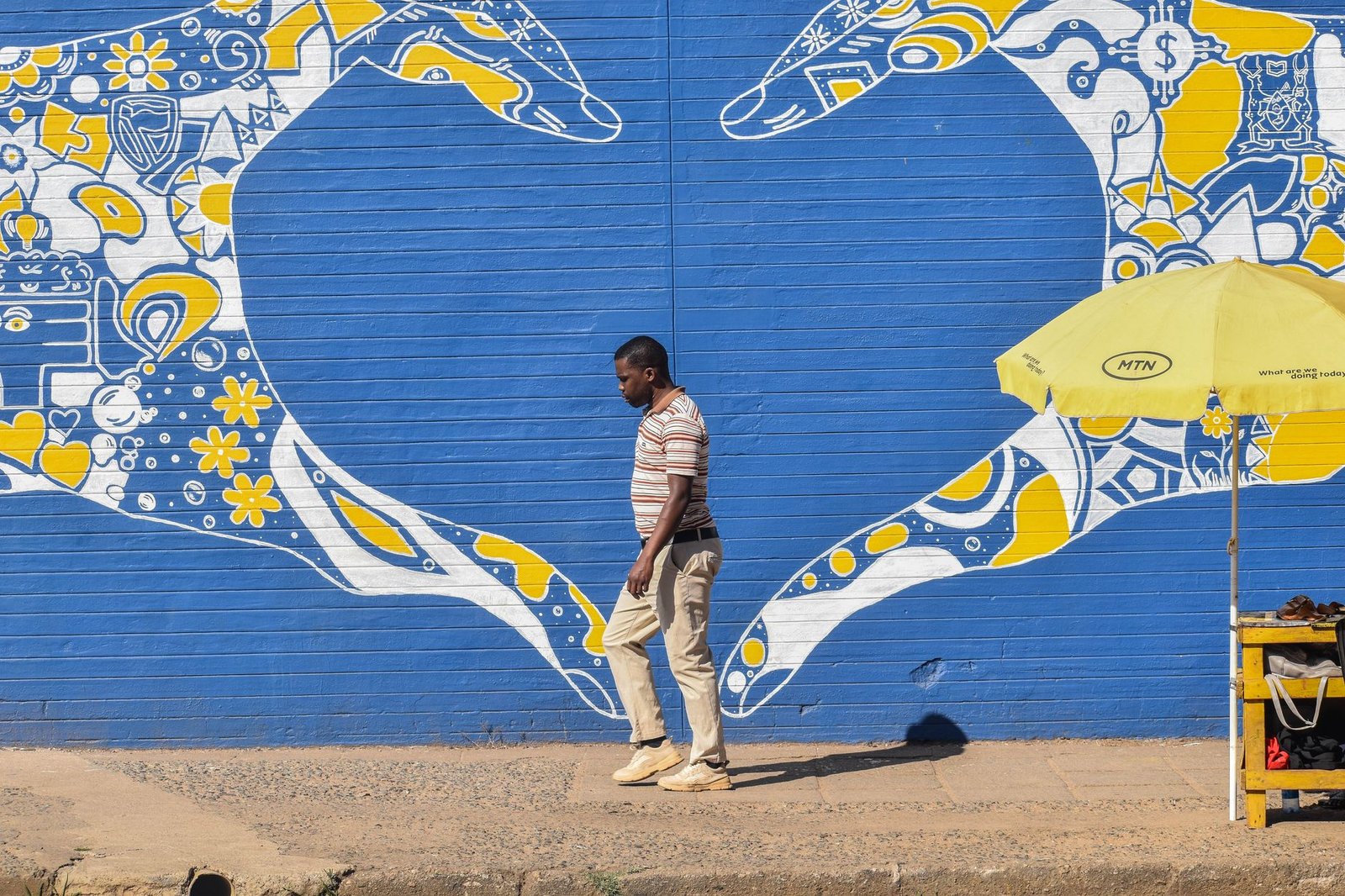 A man walks past a vibrant blue and yellow mural in Manzini, Eswatini, on a sunny day.