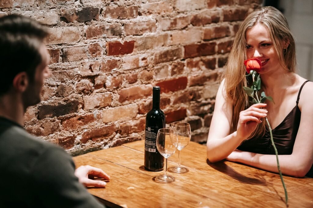 Young couple in elegant clothes sitting at wooden table near brick wall in light restaurant while enjoying date with red rose