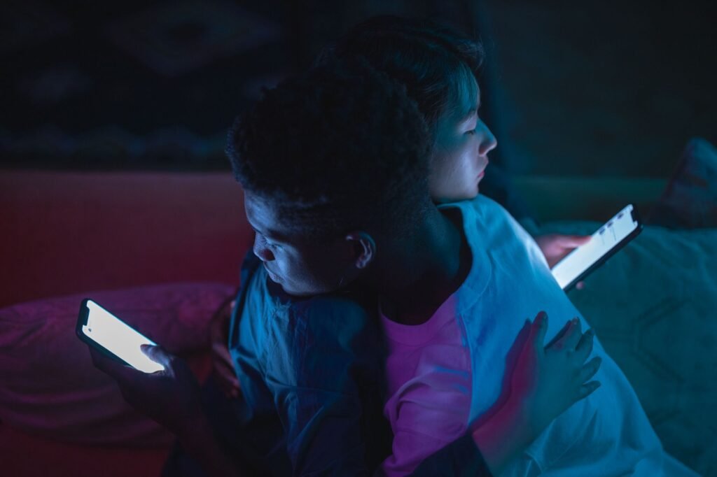 Interracial couple embracing while using smartphones, casting a warm glow in a dark room.