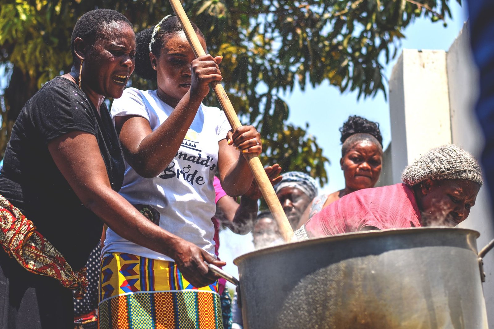 A group of women collaborating to stir a huge pot of food outdoors. Vibrant and community-focused.