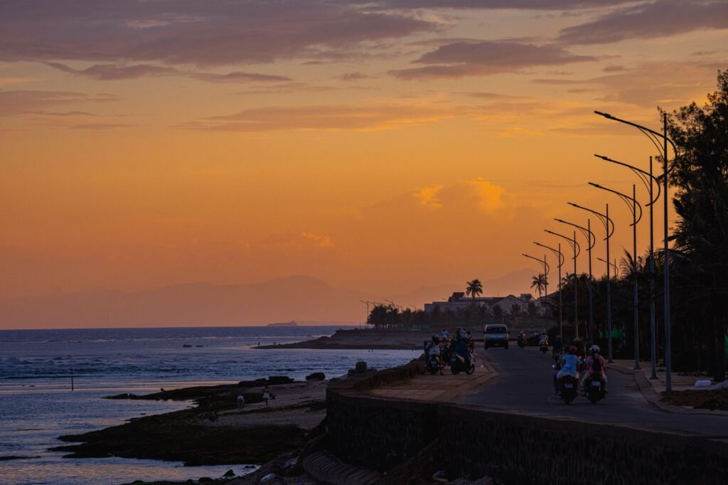 Motorcyclists ride along a coastal road at sunset with palm trees and ocean views.