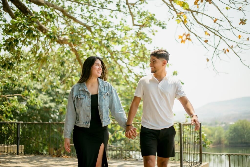 Young couple walking and holding hands in a sunny outdoor park under leafy trees.