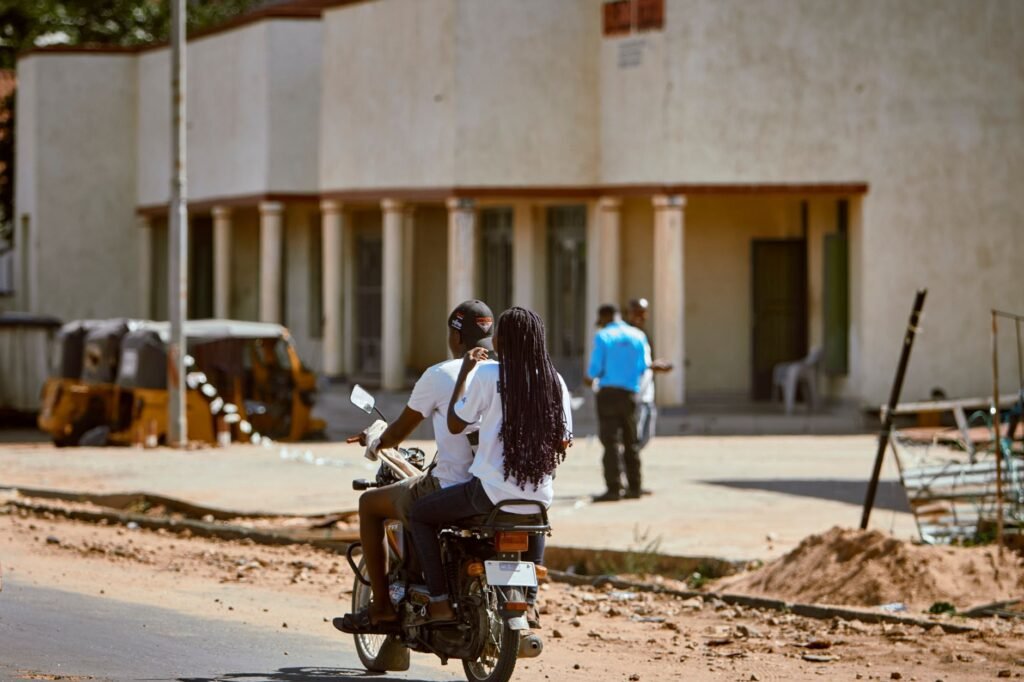A couple rides a motorcycle through a street, with city life in the backdrop.