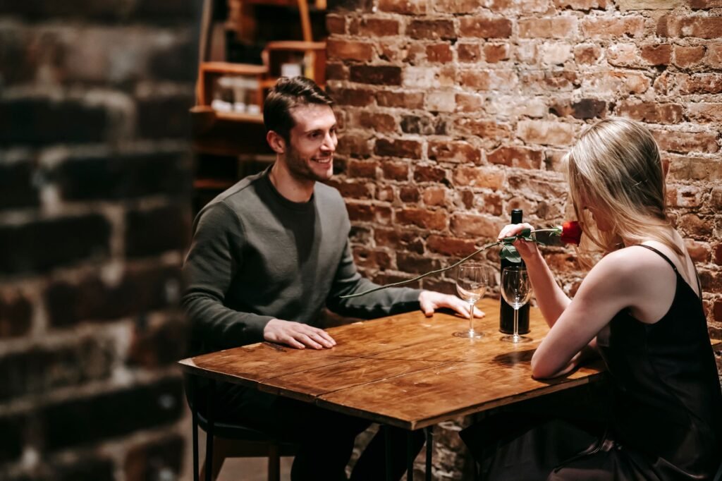 Young couple in elegant clothes sitting in light cafeteria at wooden table near brick wall and having romantic dinner with re