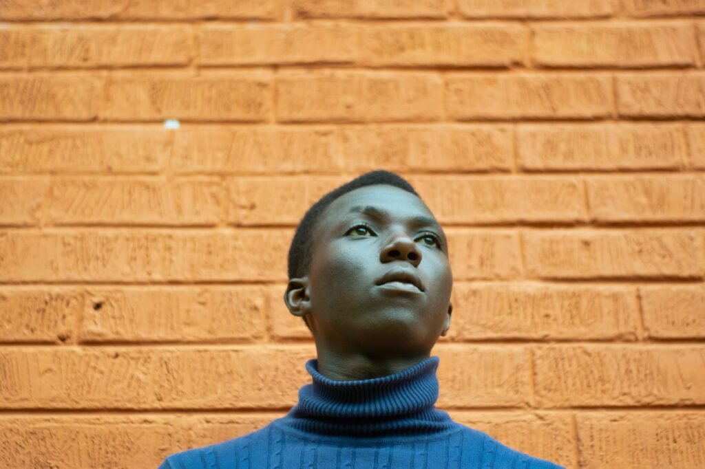 A young man in a blue turtleneck gazing thoughtfully against an orange brick wall.