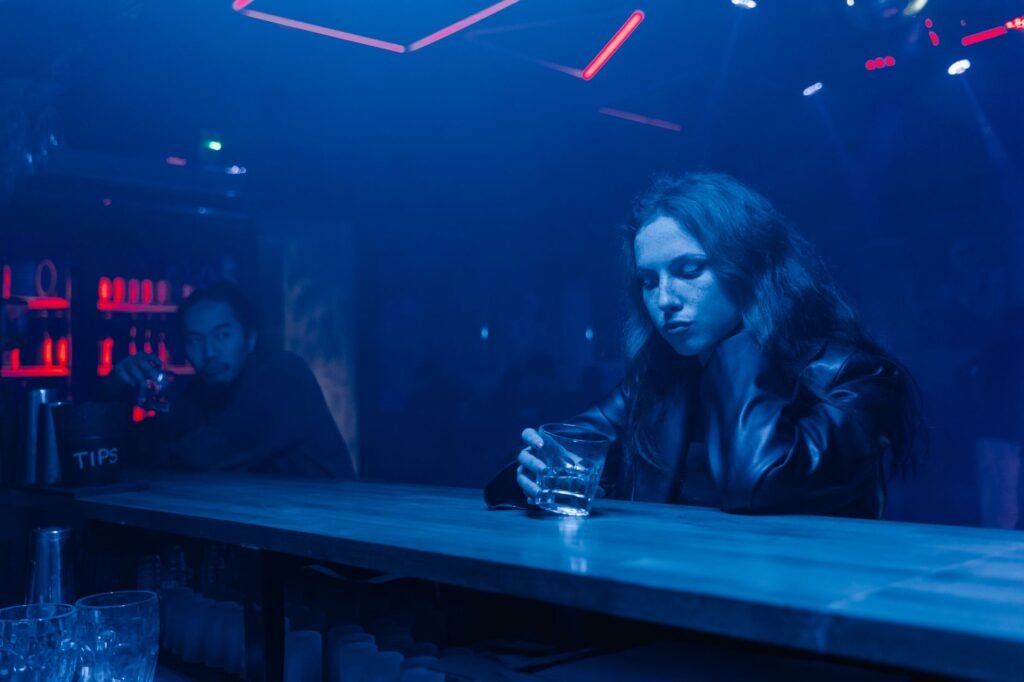A contemplative woman sits with a drink in a dimly lit bar, surrounded by blue neon lights.