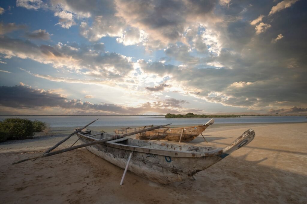 A weathered fishing boat rests on a sandy beach during a dramatic sunset, overlooking the ocean horizon.