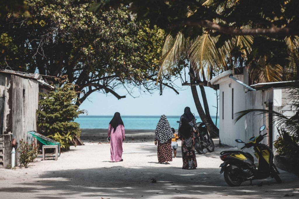A group of women in hijabs walking with a child on a coastal pathway lined with trees and scooters.