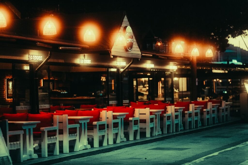 Illuminated outdoor restaurant with empty tables and chairs at evening, adorned with warm lights.