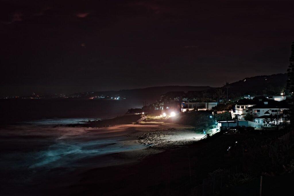 A peaceful night scene capturing the illuminated beach and coastal buildings, with waves gently lapping the shore.