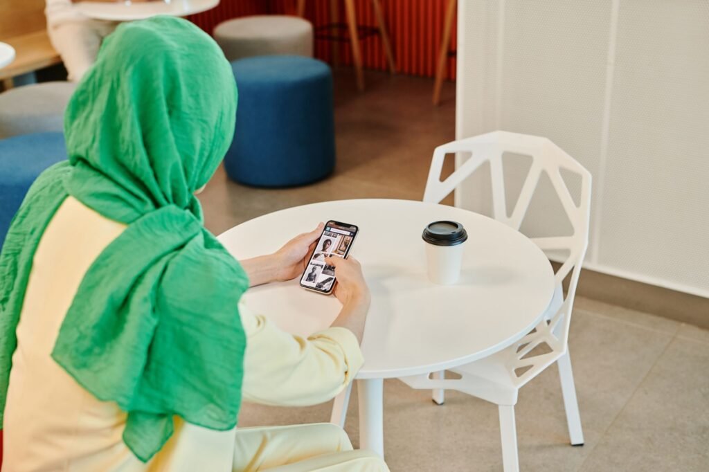 A woman wearing a green hijab is scrolling on her smartphone while sitting at a cafe table.