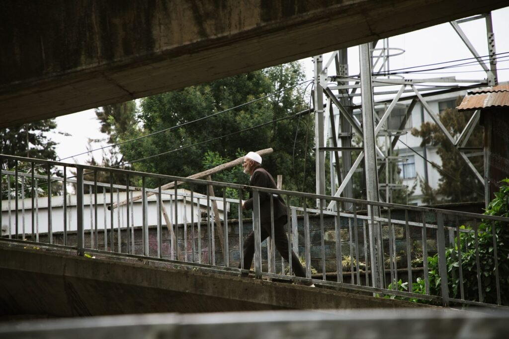 Elderly man with cane walks on an overpass in a bustling urban area, surrounded by greenery.