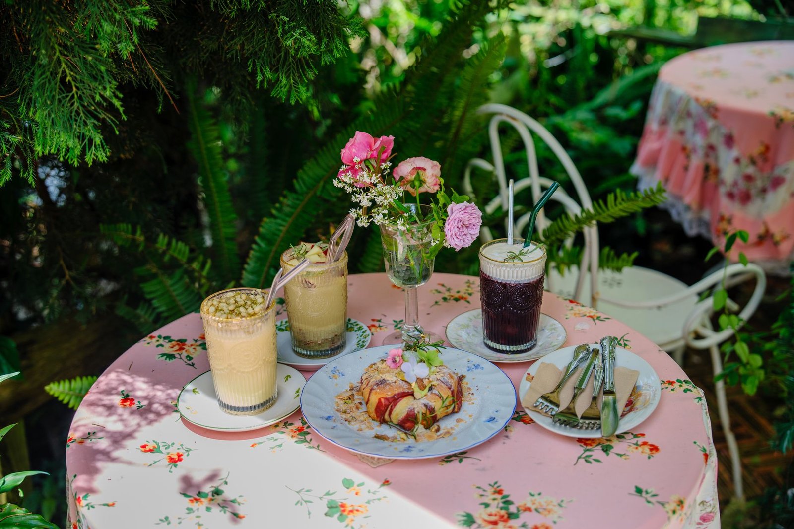 Cozy outdoor cafe setting with floral decor, pastries, and beverages on a sunny day garden table.