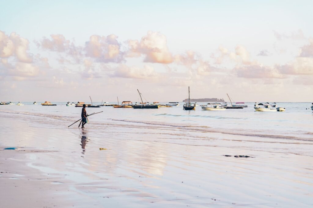 A peaceful beach with a lone person walking and boats on the horizon at sunrise.