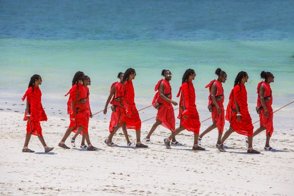 Maasai warriors in traditional attire walking along a pristine tropical beach with turquoise waters.