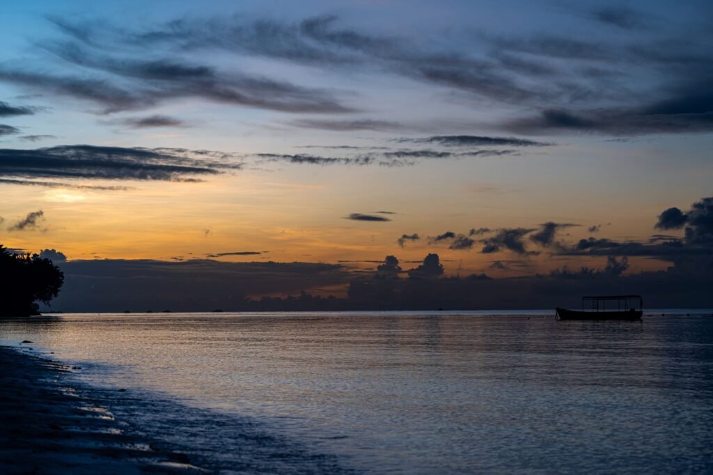 A tranquil beach sunset with a silhouetted boat on calm waters, perfect for serene landscapes.