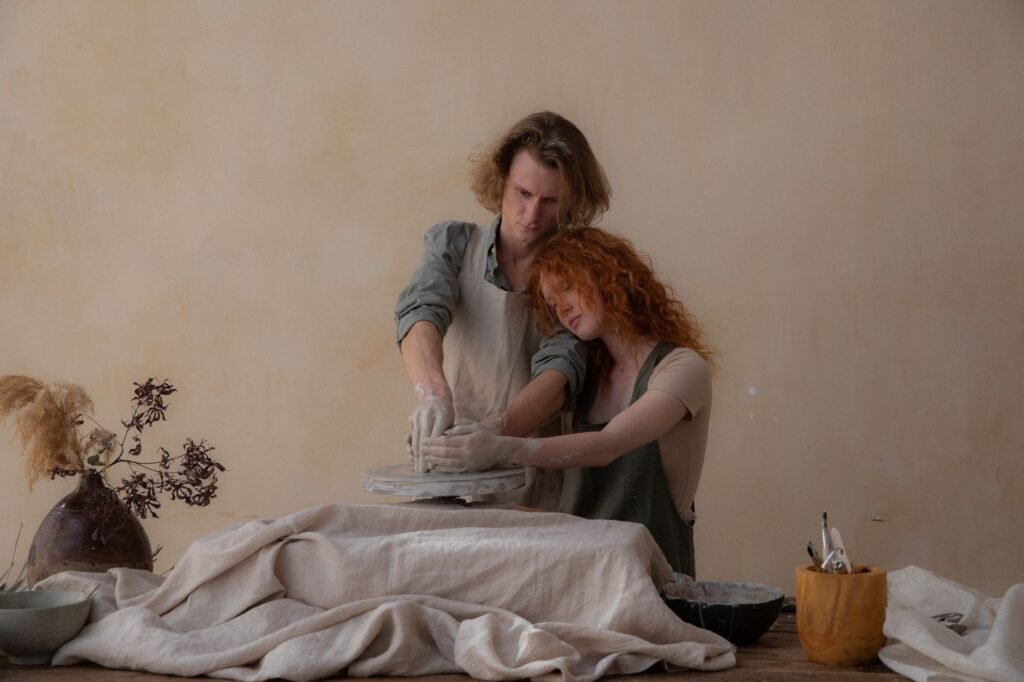 Loving couple in aprons standing near table with supplies while creating clay bowls on wheel