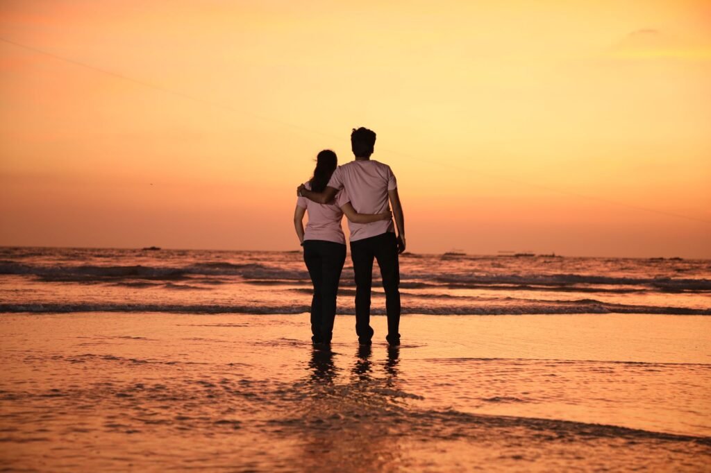 A couple stands embracing on the beach during a warm sunset, enjoying a romantic moment.