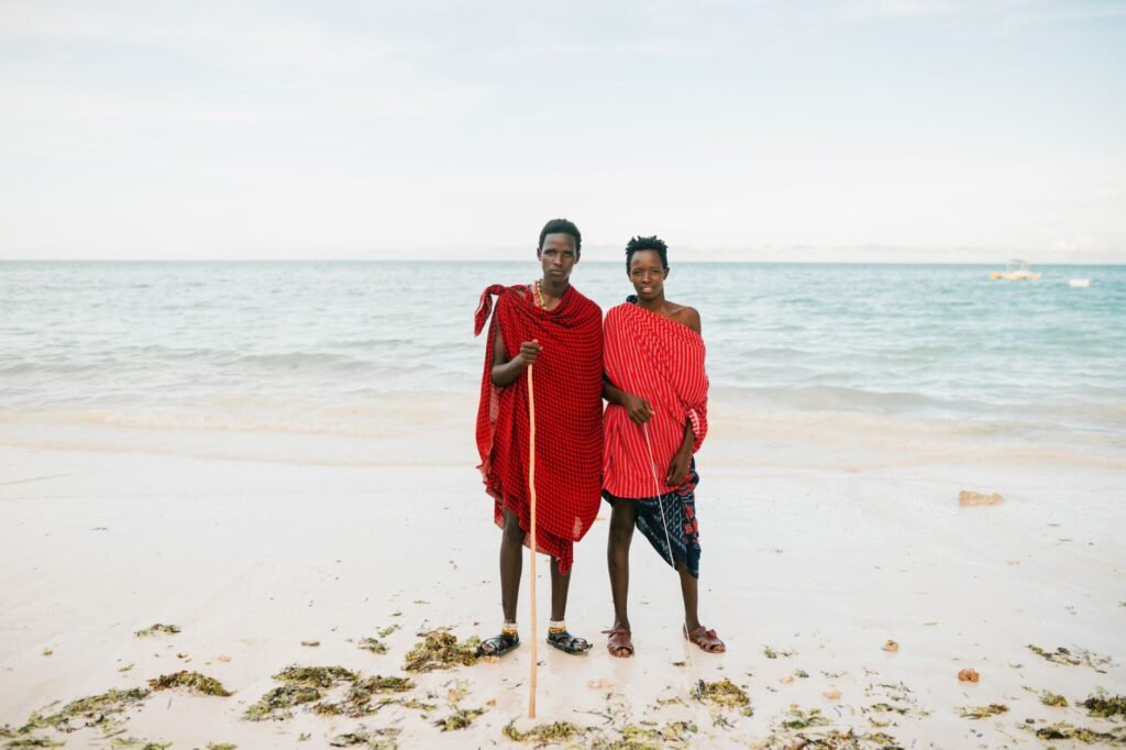 African father and teenage son in traditional red clothes standing on sandy beach and looking at camera