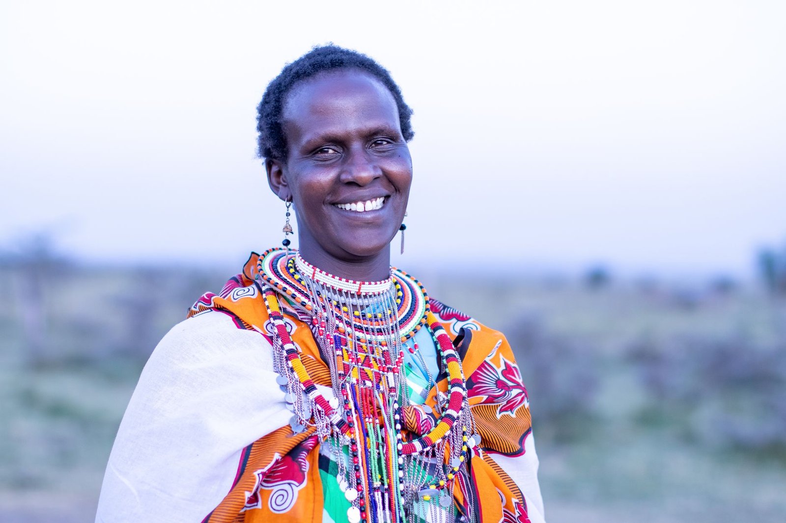 A smiling Maasai woman wearing colorful traditional attire and jewelry, captured outdoors in Kenya.