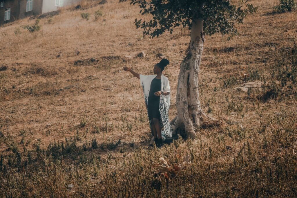 A woman stands beside a tree on a dry hillside, capturing a serene moment outdoors.