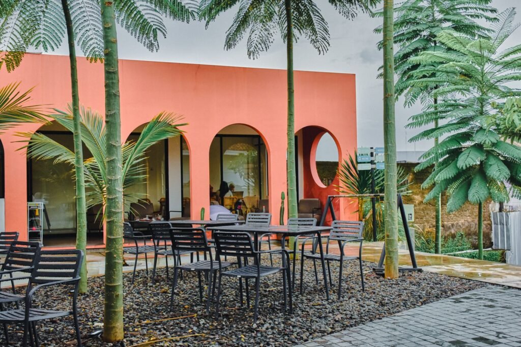 Outdoor seating at a vibrant cafe with tropical palm trees, featuring a coral-colored facade with arched windows.