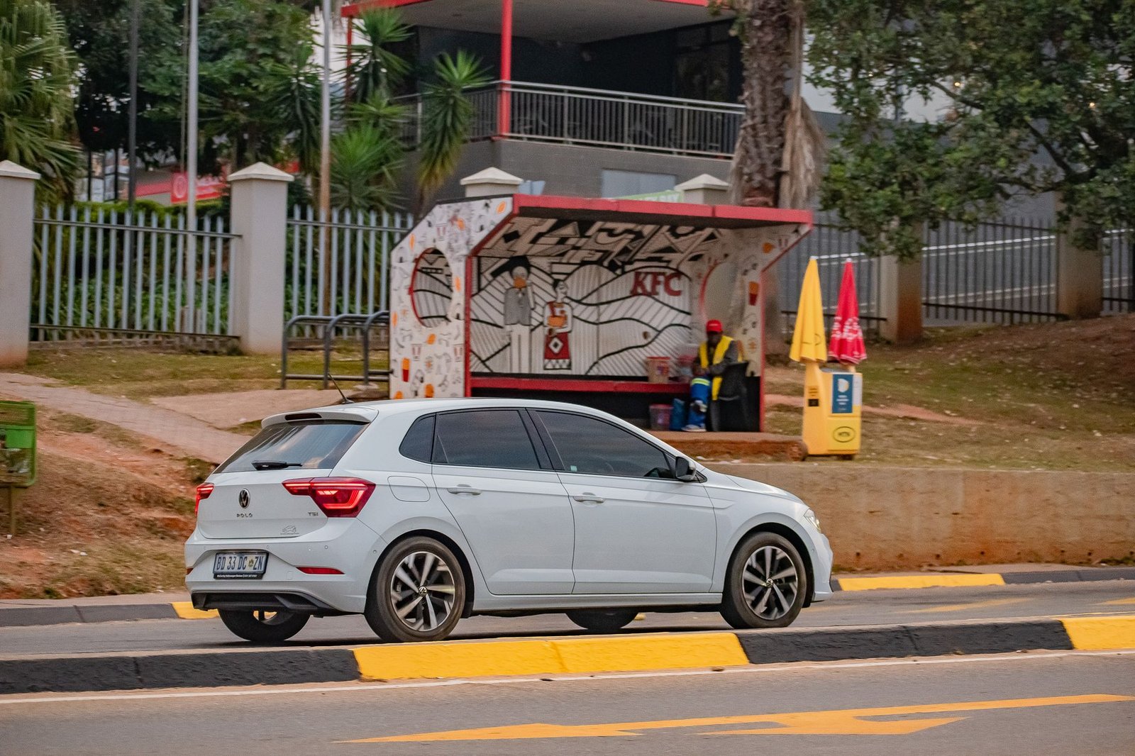 A car passes a colorful bus stop with a mural in Malkerns, Eswatini, featuring a person seated nearby.