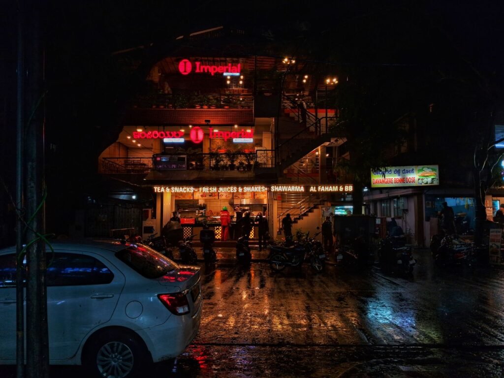 Rainy night street scene with cafes in Koramangala, India, capturing vibrant urban life.