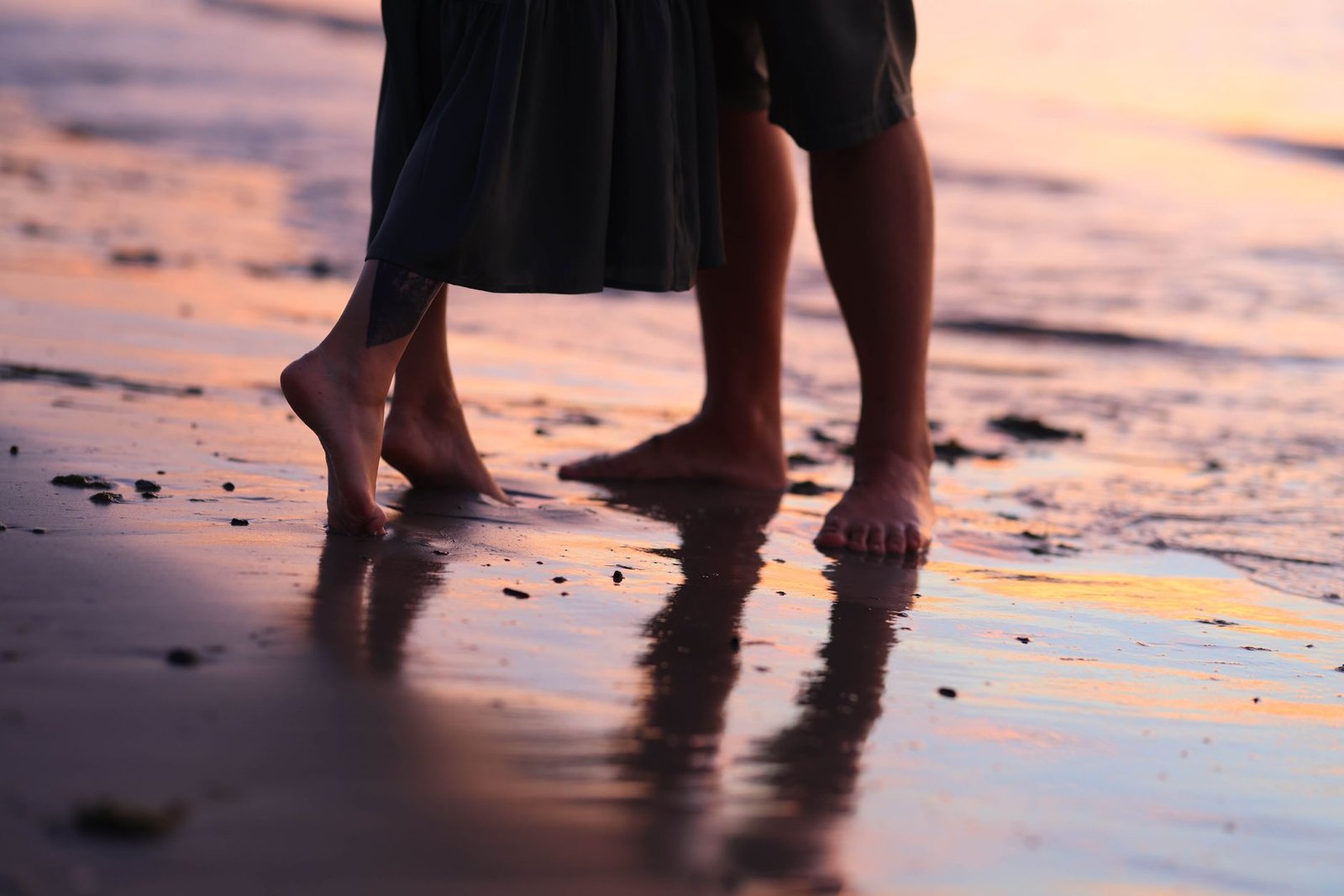A couple stands barefoot on a beach at sunset, creating serene reflections in the wet sand.