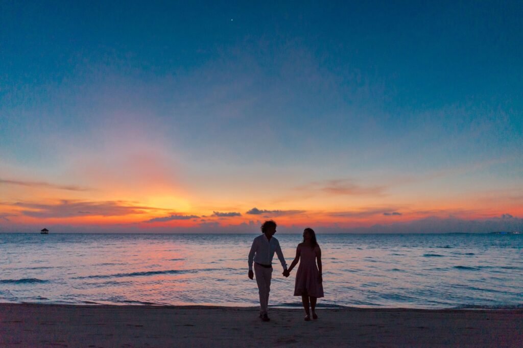 A couple holding hands walking along a beach at sunset, creating a silhouette against a vibrant sky.