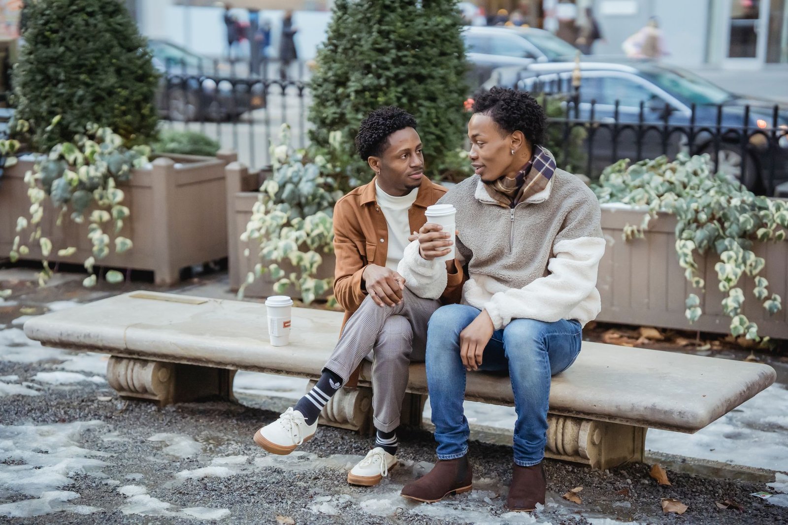 Full body of African American male couple with takeaway coffee looking at each other while sitting on bench on snowy street