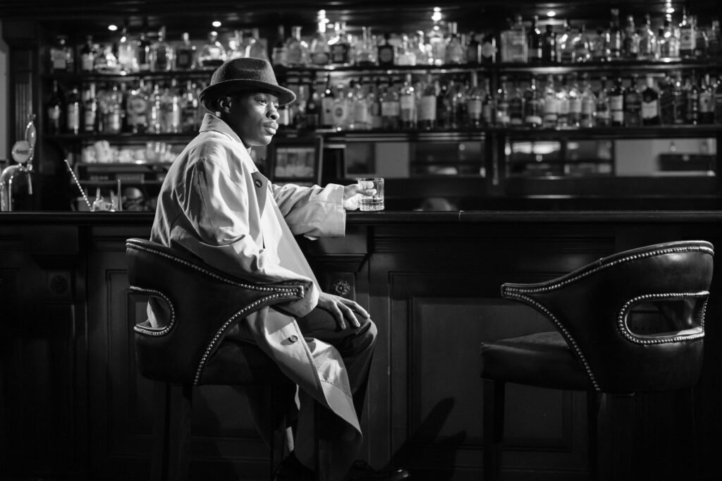 Black and white noir-style photo of a man with a fedora sitting at a bar with a drink.