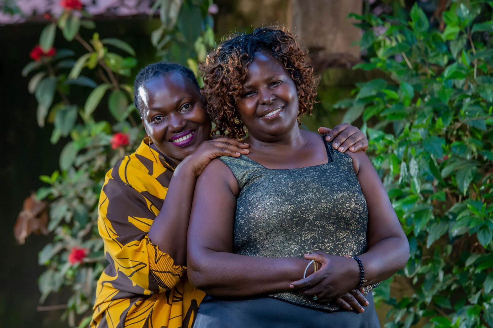 Two smiling women embracing amidst vibrant greenery outdoors, displaying warmth and friendship.