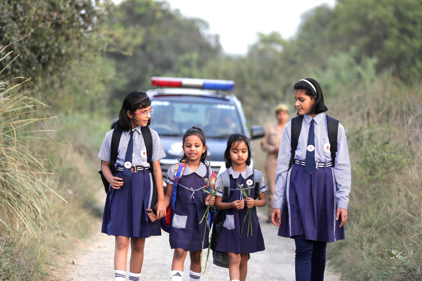 Four young school girls walking together in rural area, escorted by a police car.