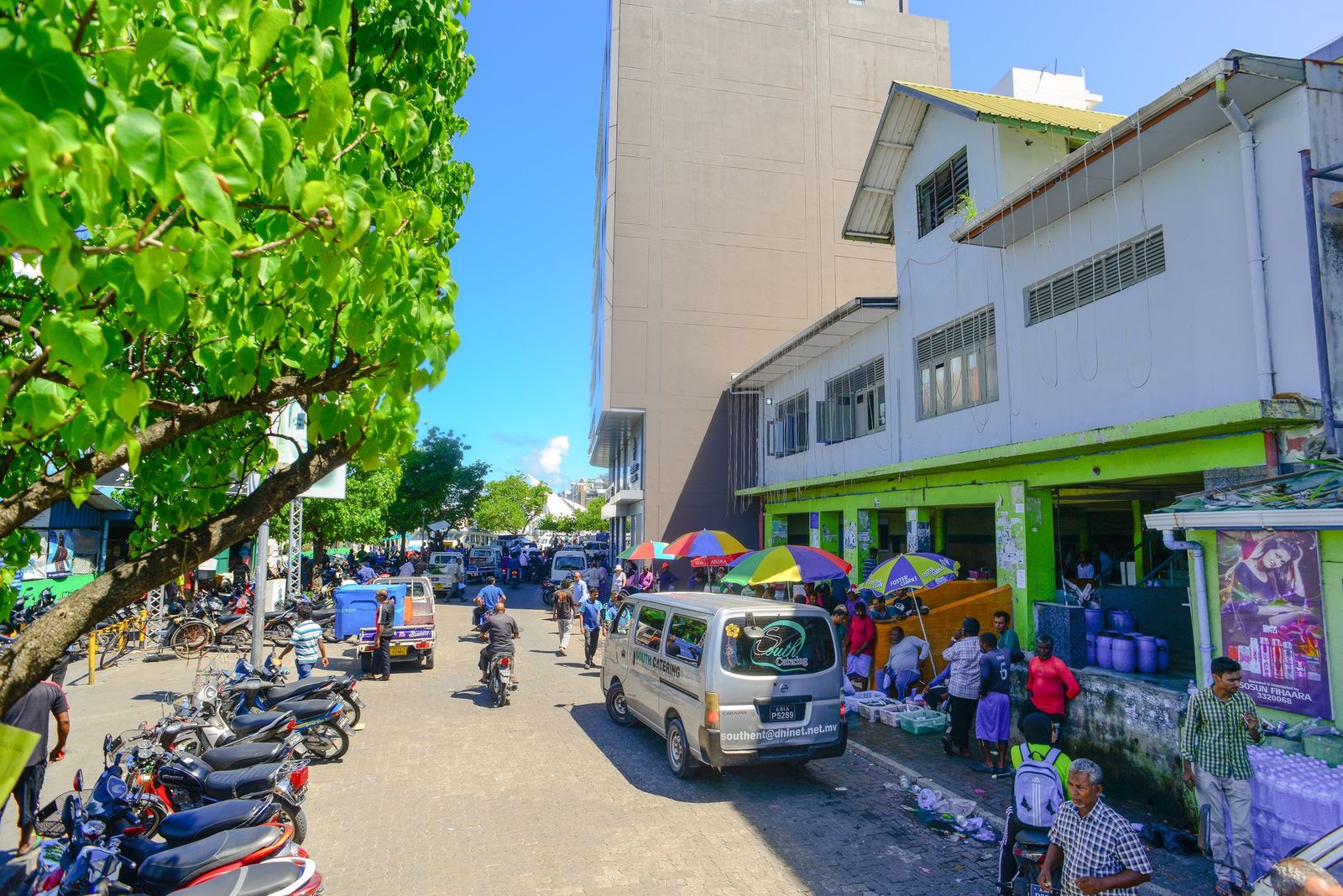 A vibrant street with market stalls, people, and vehicles in an urban setting under a sunny sky.