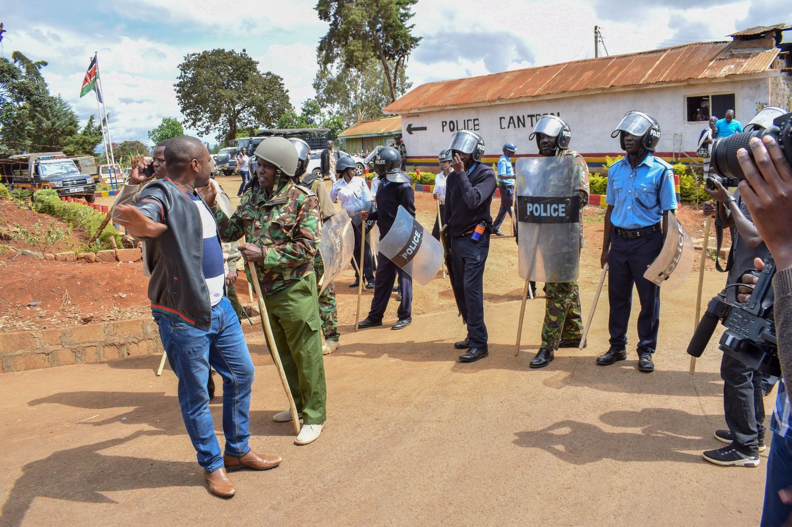 Police officers equipped with shields face a protester in a rural area.