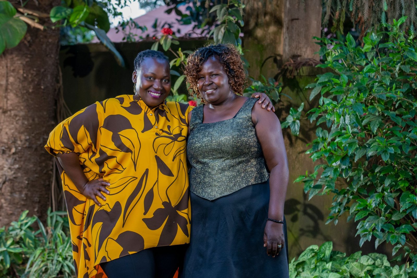 Two women smiling and embracing in a lush garden, wearing vibrant clothing.